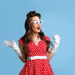 Kobieta w czerwonej sukience w białe grochy, białych rękawiczkach i okularach w stylu pin-up girl stylizacja, z uniesionymi rękami.