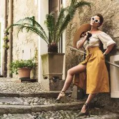 Kobieta w letniej stylizacji z espadrylami na koturnie, oparta o kamienną ścianę, z kapeluszem w dłoni.