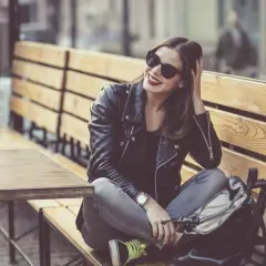 Dziewczyna w skórzanej kurtce i okularach przeciwsłonecznych, z czerwoną szminką, siedzi na ławce w kawiarni. Jej styl to rockowy kobiecy.