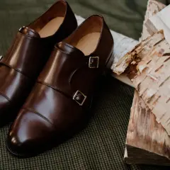 Eleganckie brązowe buty do czarnych spodni, z podwójnymi klamrami, leżą obok polan brzozowych na tweedowym tle.