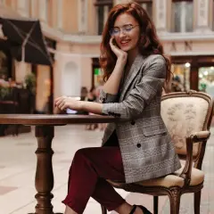 Elegancka kobieta w bordowych spodniach i marynarce w kratę, siedząca przy stoliku. Stylowe bordowe spodnie dodają jej szyku.