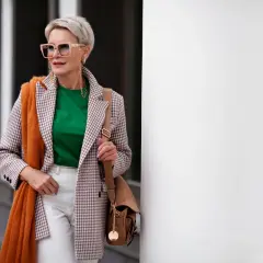 Elegancka kobieta po 60. prezentuje modne stylizacje: marynarka w kratkę, zielona bluzka, białe spodnie, pomarańczowy szal i torebka.