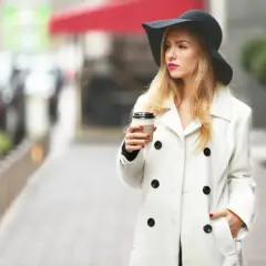 Elegancka kobieta w kapeluszu i białym płaszczu, z kawą w ręku, uosabia paryską modę.