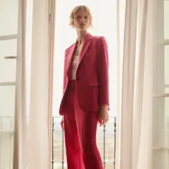 Elegancka stylizacja na chrzciny ze spodniami: kobieta w różowym garniturze stoi przy oknie.