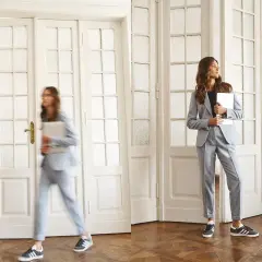 Dwie kobiety w eleganckich, jasnoszarych garniturach, które idealnie wpisują się w **casual dress code**. Jedna spieszy się z laptopem, druga czeka, trzymając tablet.