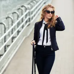 Eleganckie stylizacje z czarnymi spodniami: kobieta w ciemnej kurtce, białej koszuli z kokardą i okularach przeciwsłonecznych idzie po moście.