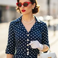 Elegancka kobieta w granatowej sukience w groszki, białych rękawiczkach i czerwonych okularach przeciwsłonecznych. Stylizacje retro.