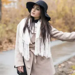 Elegancka kobieta w beżowym płaszczu i czarnym kapeluszu, z białym szalem, zastanawia się, jaki szal do beżowego płaszcza będzie najlepszy.