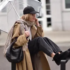 Kobieta w beżowym płaszczu, czapce z daszkiem i jeansach prezentuje modne emu stylizacje, chroniąc się pod przezroczystym parasolem.
