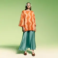 Modelka w okularach w kształcie serc i pomarańczowym swetrze z falistym wzorem. Stylizacja w stylu color block z turkusową, lejącą spódnicą.