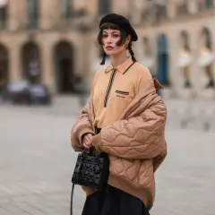 Dziewczyna w czarnym beretcie, z warkoczykami, w beżowej kurtce i bluzce. Pokazuje, jak założyć beret, by wyglądać stylowo.