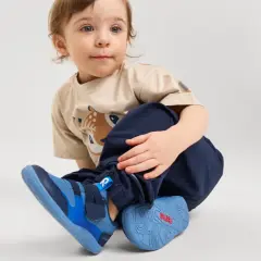 Małe dziecko w beżowej koszulce z jelonkiem i granatowych spodniach, nosi niebieskie buty sportowe.