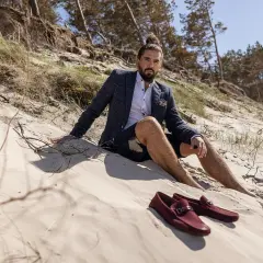 Mokasyny męskie stylizacje na plaży. Mężczyzna w marynarce i szortach siedzi na piasku, obok niego leżą bordowe mokasyny.