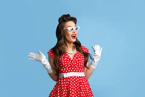Kobieta w czerwonej sukience w białe grochy, białych rękawiczkach i okularach w stylu pin-up girl stylizacja, z uniesionymi rękami.