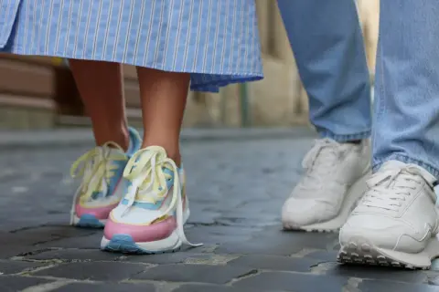 Kolorowe, modne sneakersy 2026 na nogach osoby w sukience i białe na nogach w jeansach, spacerujące po bruku.