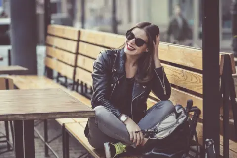 Dziewczyna w skórzanej kurtce i okularach przeciwsłonecznych, z czerwoną szminką, siedzi na ławce w kawiarni. Jej styl to rockowy kobiecy.