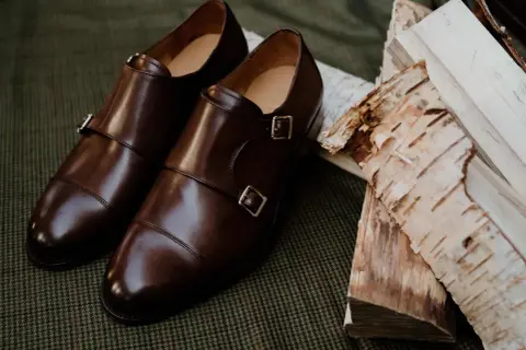 Eleganckie brązowe buty do czarnych spodni, z podwójnymi klamrami, leżą obok polan brzozowych na tweedowym tle.