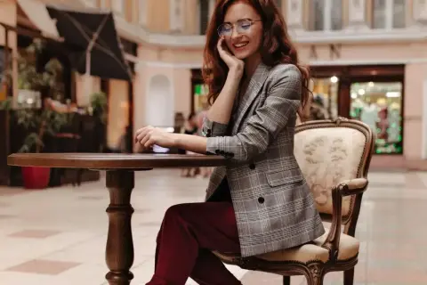 Elegancka kobieta w bordowych spodniach i marynarce w kratę, siedząca przy stoliku. Stylowe bordowe spodnie dodają jej szyku.