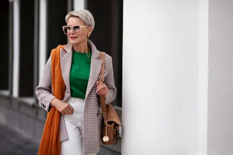 Elegancka kobieta po 60. prezentuje modne stylizacje: marynarka w kratkę, zielona bluzka, białe spodnie, pomarańczowy szal i torebka.