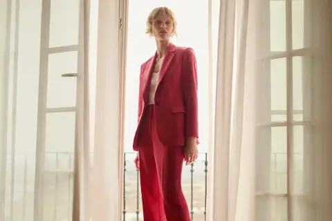 Elegancka stylizacja na chrzciny ze spodniami: kobieta w różowym garniturze stoi przy oknie.