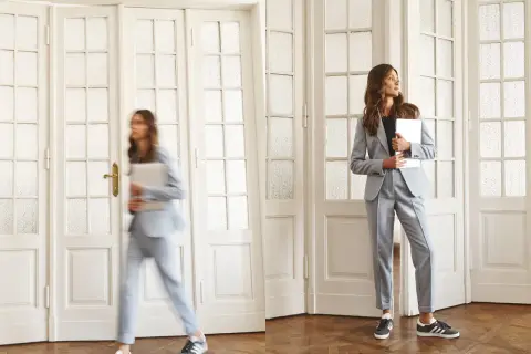 Dwie kobiety w eleganckich, jasnoszarych garniturach, które idealnie wpisują się w **casual dress code**. Jedna spieszy się z laptopem, druga czeka, trzymając tablet.