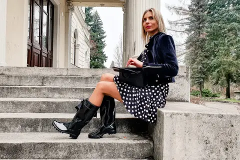 Elegancka kobieta w grochy, w czarnych, błyszczących kaloszach ze skarpetą, siedzi na kamiennych schodach.