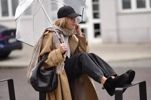 Kobieta w beżowym płaszczu, czapce z daszkiem i jeansach prezentuje modne emu stylizacje, chroniąc się pod przezroczystym parasolem.