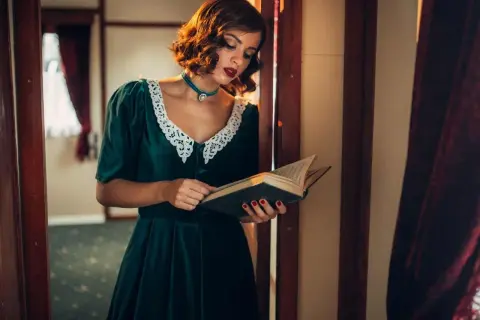 Elegancka kobieta w sukni z lat 40. czyta książkę. Jej stylizacja, z koronkowym kołnierzykiem i biżuterią, idealnie oddaje modę tamtych lat.