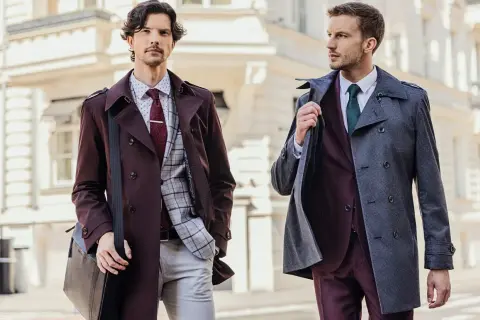 Dwóch eleganckich mężczyzn w garniturach. Jeden w bordowym płaszczu i brązowych butach do czarnego garnituru, drugi w szarym płaszczu.