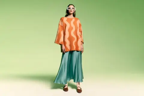 Modelka w okularach w kształcie serc i pomarańczowym swetrze z falistym wzorem. Stylizacja w stylu color block z turkusową, lejącą spódnicą.