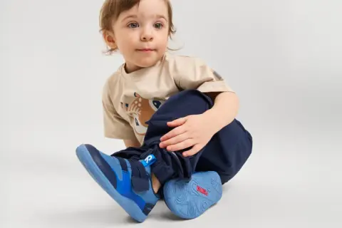 Małe dziecko w beżowej koszulce z jelonkiem i granatowych spodniach, nosi niebieskie buty sportowe.