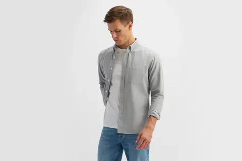 Młody mężczyzna w eleganckim ubiorze męskim: szara koszula z długim rękawem, biały t-shirt i jeansy.
