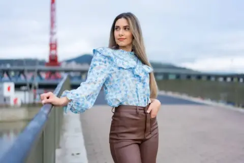 Kobieca, błękitna bluzka w kwiaty z falbankami idealnie pasuje do brązowych, skórzanych spodni.