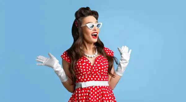Kobieta w czerwonej sukience w białe grochy, białych rękawiczkach i okularach w stylu pin-up girl stylizacja, z uniesionymi rękami.