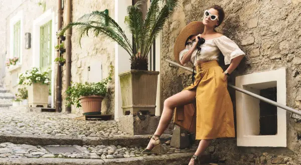 Kobieta w letniej stylizacji z espadrylami na koturnie, oparta o kamienną ścianę, z kapeluszem w dłoni.