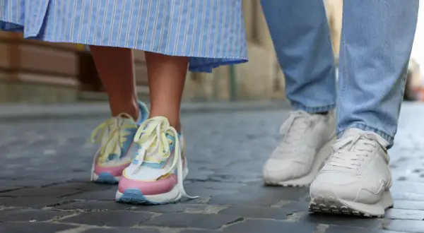 Kolorowe, modne sneakersy 2026 na nogach osoby w sukience i białe na nogach w jeansach, spacerujące po bruku.