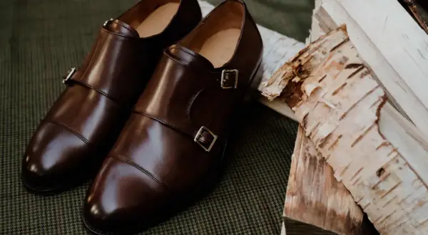 Eleganckie brązowe buty do czarnych spodni, z podwójnymi klamrami, leżą obok polan brzozowych na tweedowym tle.