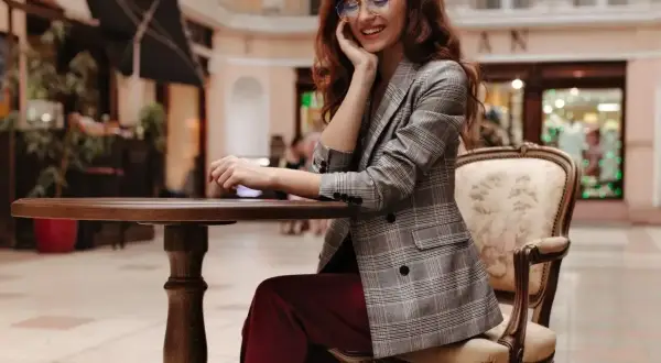 Elegancka kobieta w bordowych spodniach i marynarce w kratę, siedząca przy stoliku. Stylowe bordowe spodnie dodają jej szyku.