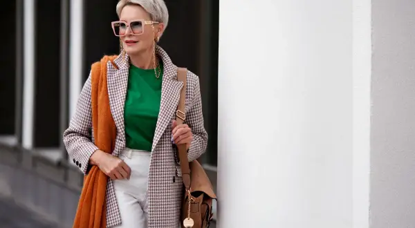 Elegancka kobieta po 60. prezentuje modne stylizacje: marynarka w kratkę, zielona bluzka, białe spodnie, pomarańczowy szal i torebka.