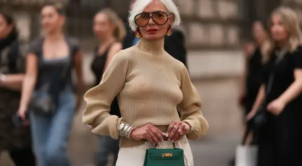 Elegancka kobieta prezentuje stylowe stylizacje dla dojrzałych pań. Beżowy golf, wzorzysta spódnica i zielona torebka tworzą szykowny look.