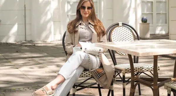 Elegancka kobieta w stylu old money damski, siedząca przy stoliku w kawiarni, w beżowym płaszczu i białych spodniach.