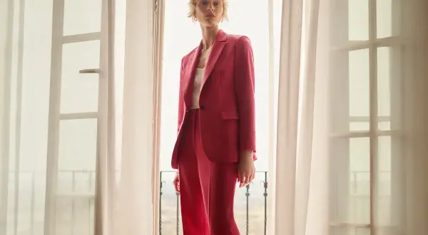 Elegancka stylizacja na chrzciny ze spodniami: kobieta w różowym garniturze stoi przy oknie.