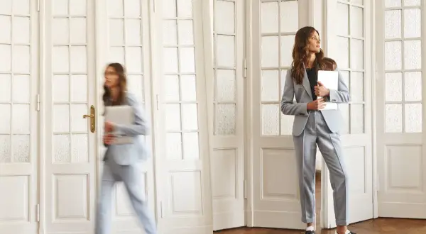 Dwie kobiety w eleganckich, jasnoszarych garniturach, które idealnie wpisują się w **casual dress code**. Jedna spieszy się z laptopem, druga czeka, trzymając tablet.