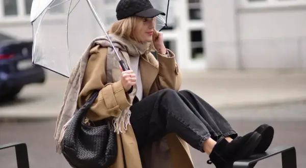 Kobieta w beżowym płaszczu, czapce z daszkiem i jeansach prezentuje modne emu stylizacje, chroniąc się pod przezroczystym parasolem.