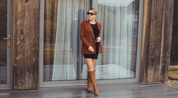 Elegancka kobieta w brązowej marynarce i czarnej sukience, prezentująca camelowe kozaki stylizacje.