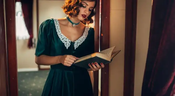Elegancka kobieta w sukni z lat 40. czyta książkę. Jej stylizacja, z koronkowym kołnierzykiem i biżuterią, idealnie oddaje modę tamtych lat.