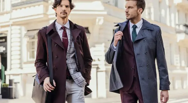 Dwóch eleganckich mężczyzn w garniturach. Jeden w bordowym płaszczu i brązowych butach do czarnego garnituru, drugi w szarym płaszczu.