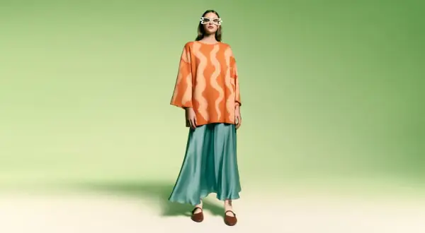 Modelka w okularach w kształcie serc i pomarańczowym swetrze z falistym wzorem. Stylizacja w stylu color block z turkusową, lejącą spódnicą.