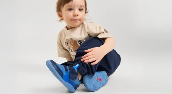 Małe dziecko w beżowej koszulce z jelonkiem i granatowych spodniach, nosi niebieskie buty sportowe.