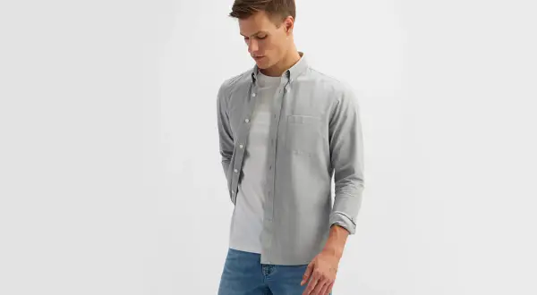 Młody mężczyzna w eleganckim ubiorze męskim: szara koszula z długim rękawem, biały t-shirt i jeansy.